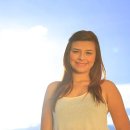 Young latin beautiful lady in her twenties with long smooth hair poses smiling and looking down to the camera for a portrait shot with the sun behind her. Backlit with the sunshining in late afternoon that causes some lens flare. Blue sky and clouds can be seen at the back.